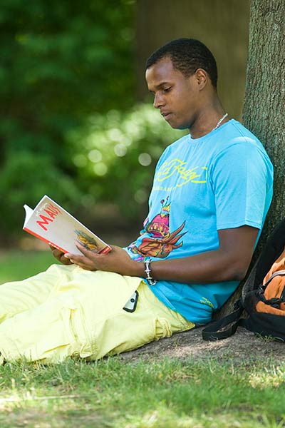 a student reading a book under the shade a tree in a grassy field